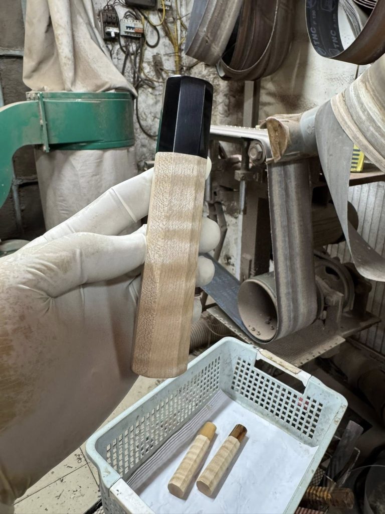 Pale figured wooden handle with a black front ferrule, held in a white glove with workshop equipment behind it.