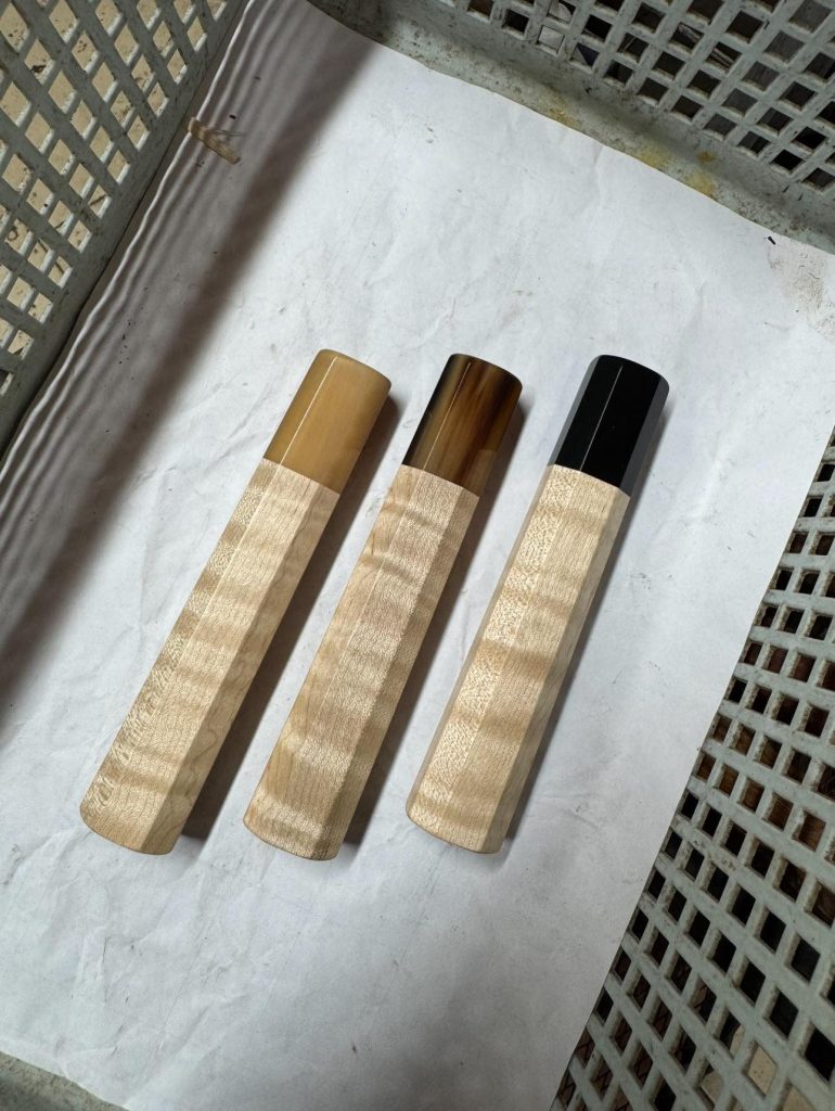 Three pale figured wooden handles with pale amber, mottled brown, and black front ferrules, arranged side by side in a basket.