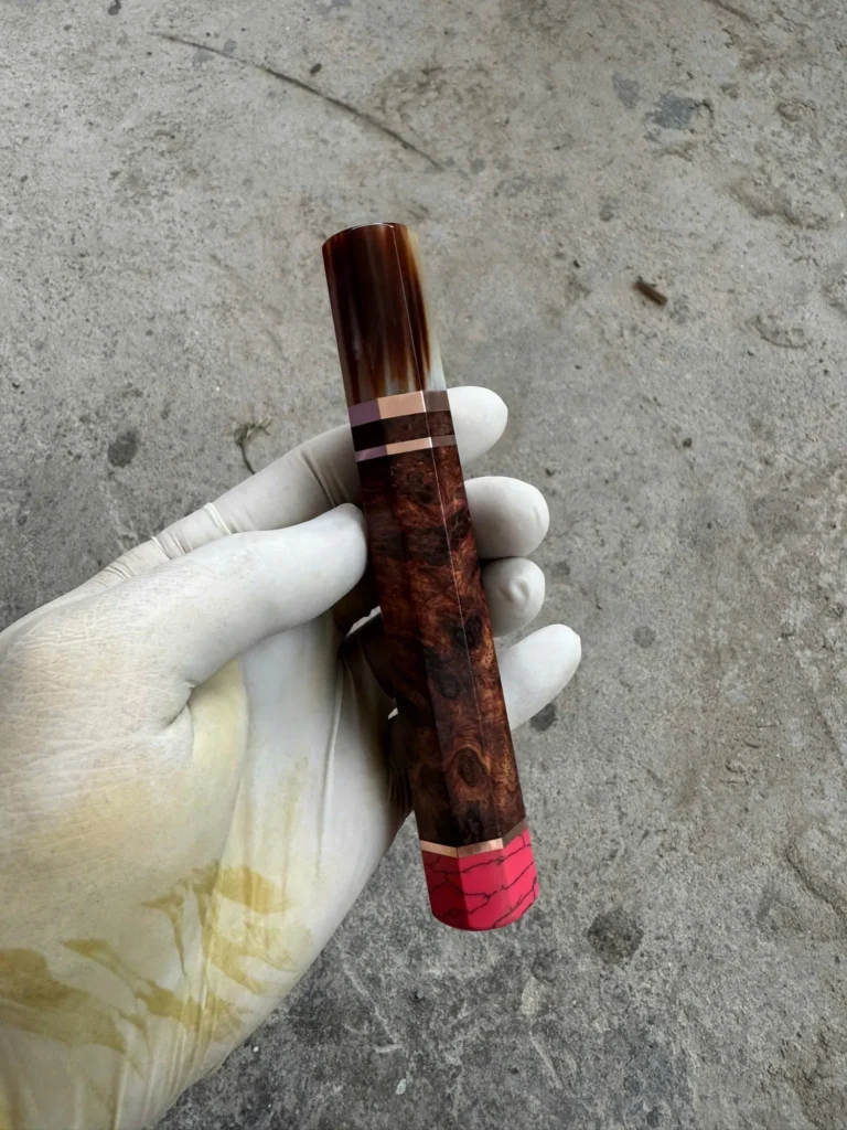 Reddish figured Amboyna burl handle with a marbled horn front ferrule, metallic spacers, and pink coral rear end cap, held in a white glove.