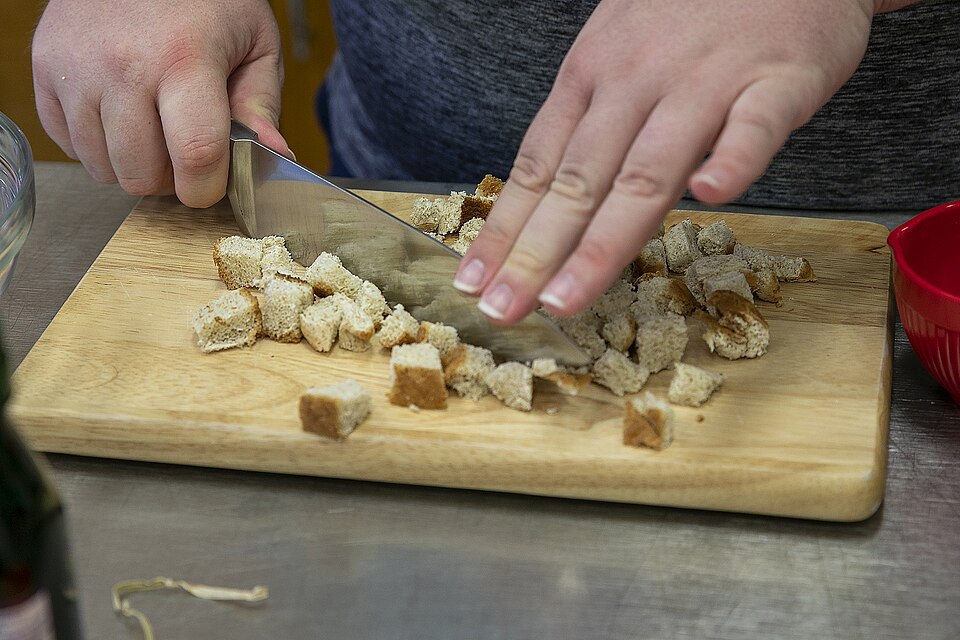 A person chops bread pieces into crumbs on a wooden cutting board.