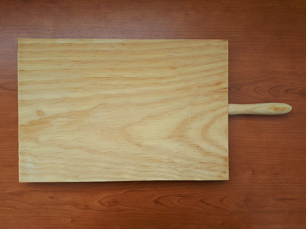 A rectangular wooden cutting board with a handle on a wooden surface.