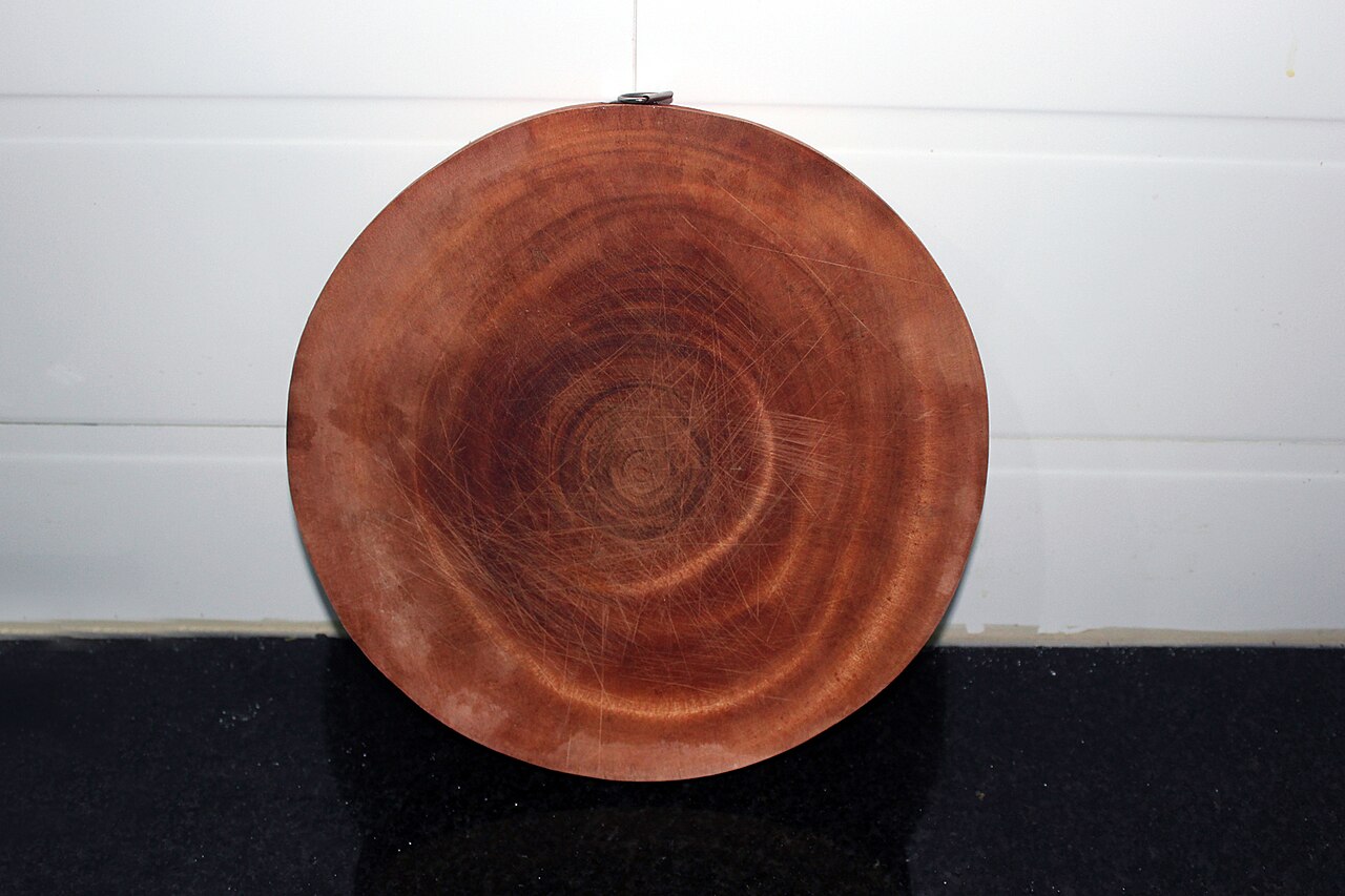 A round wooden chopping board with visible grain and knife marks, standing upright.
