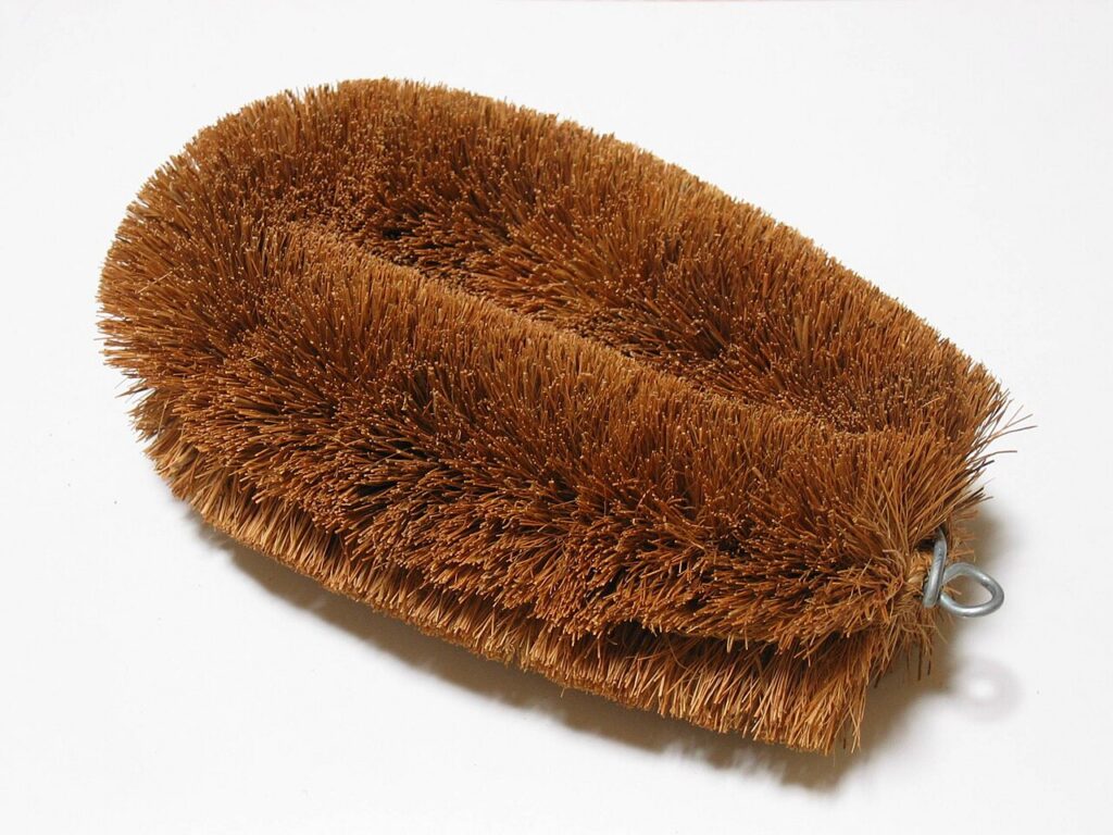 An oval brown tawashi brush with dense fibres and a small metal hanging loop.