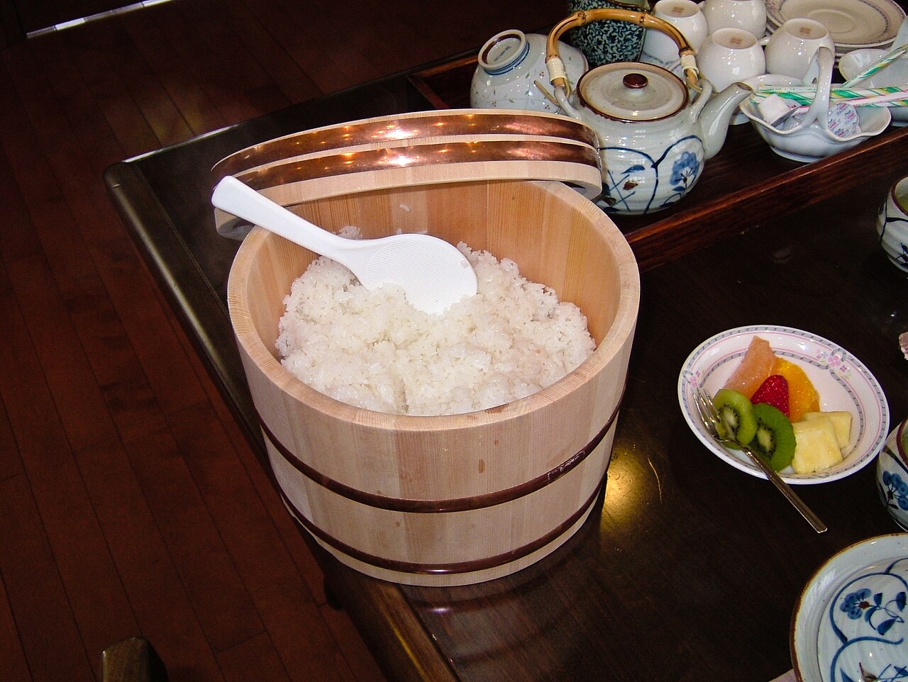 Cooked rice and a paddle inside a deeper wooden ohitsu, with its lid behind