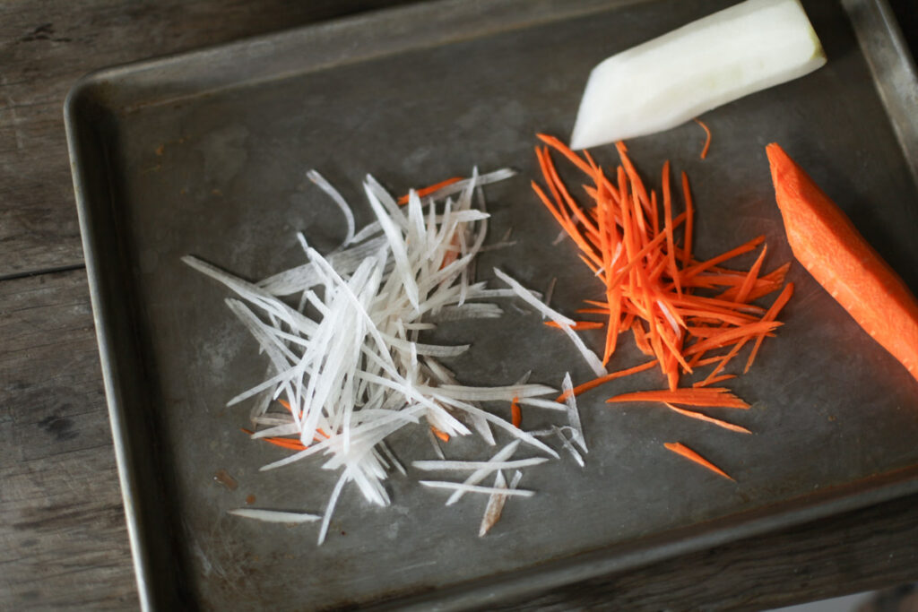 Fine strips of carrot and daikon beside larger vegetable pieces on a tray