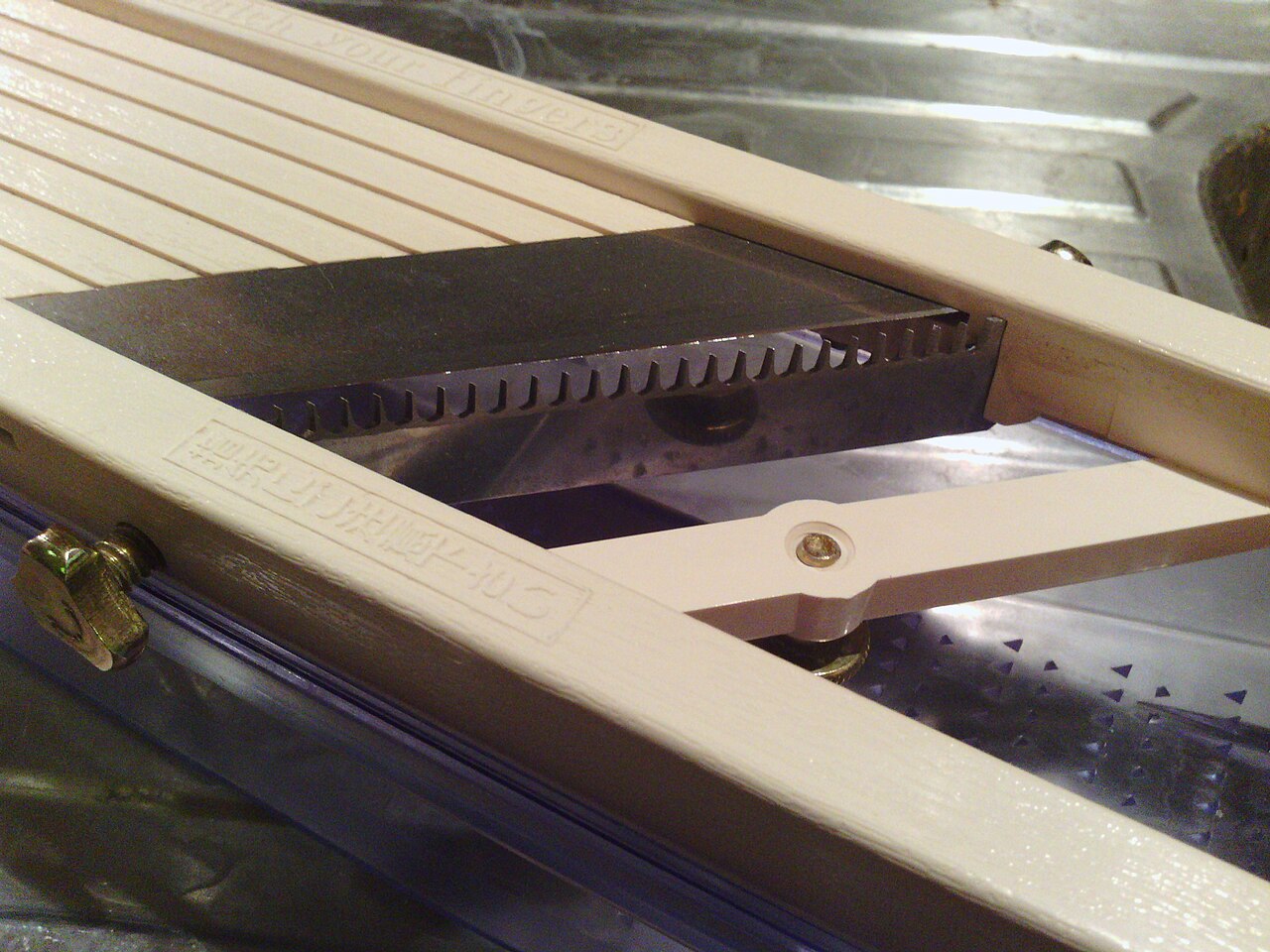 Close-up of a comb blade mounted beneath the flat blade of a Benriner slicer.