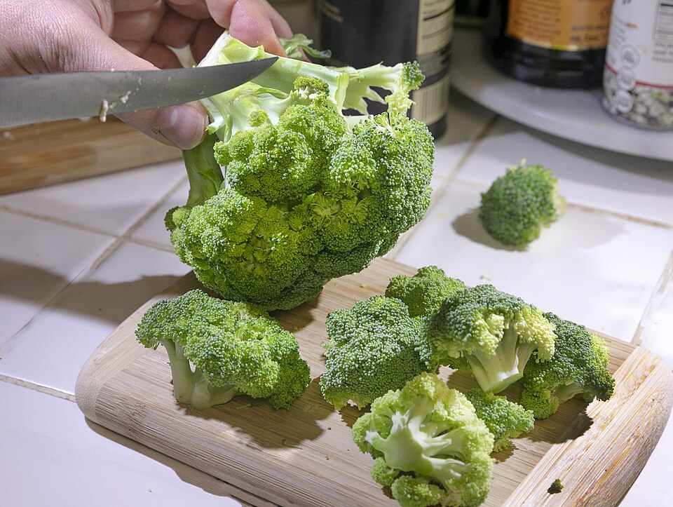A knife separates broccoli florets from the central stem on a cutting board.