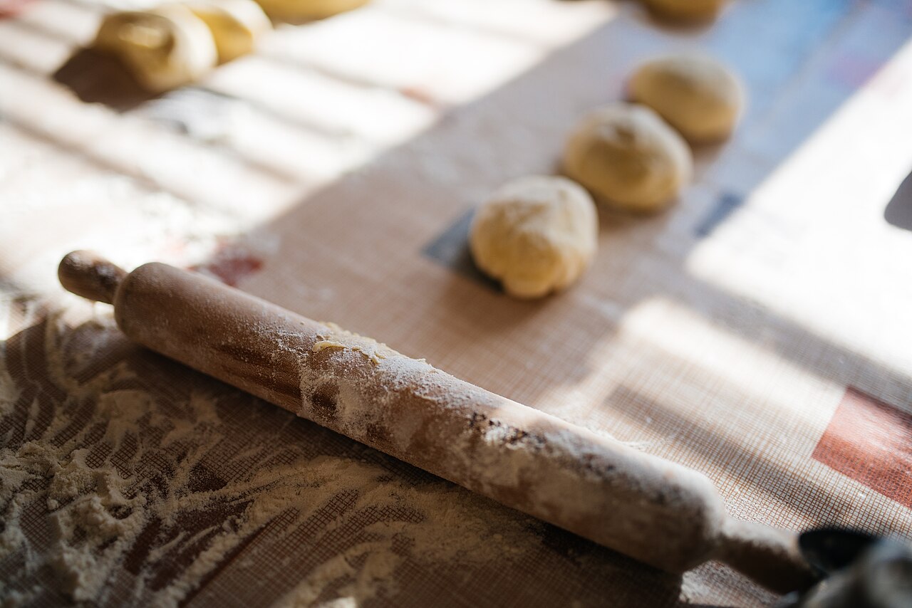A wooden rolling pin with handles lies beside portions of dough.