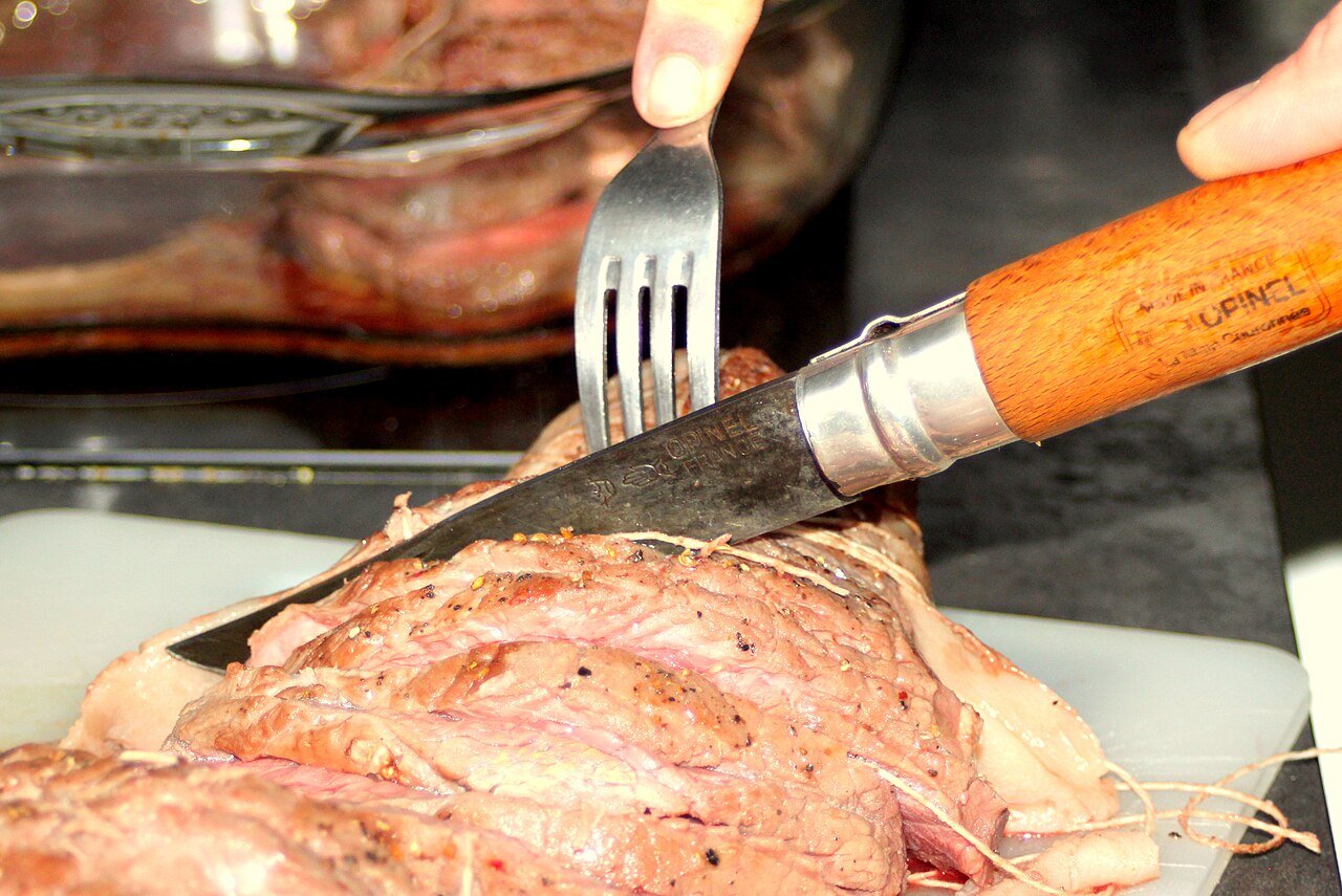 A fork holds a cooked boneless roast as a knife slices it on a cutting board.
