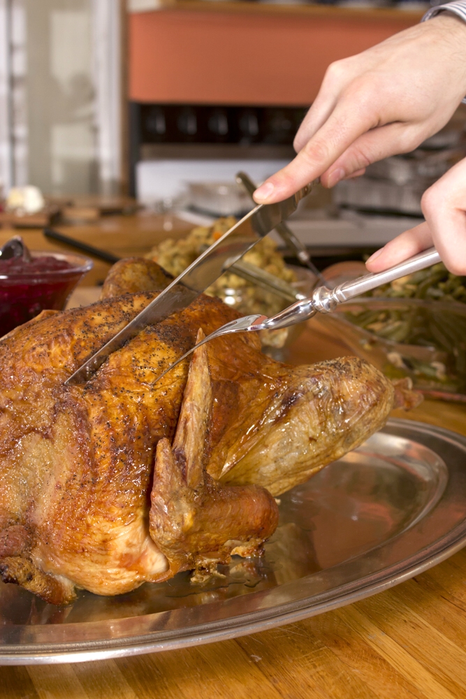 A carving fork holds a roast turkey while a knife slices meat at the table.